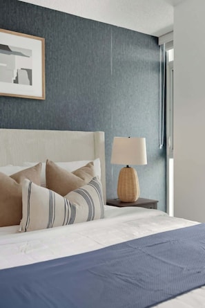 Ein ordentlich gemachtes Bett mit blauer Bettdecke und gestreifter Kopfkissen, eine Nachttischlampe auf einem Holznachttisch und ein gerahmtes Bild an der Wand.