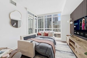 A modern bedroom with a large window, a bed with multiple pillows, a bedside table, and a TV mounted on the wall.