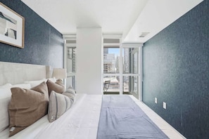 A modern hotel room with a large bed, a view of the city through a large window, and a dark-colored accent wall. - Sleek 2BR Waterfront Condo Perfect City Getaway | Parking Available (Toronto)