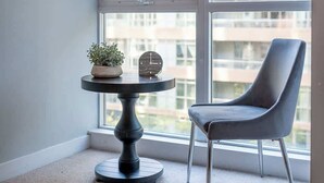 Ein Eckzimmer mit großem Fenster, einem runden Tisch mit Uhr und Pflanze sowie einem modernen Sessel.