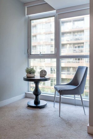 Ein Eckzimmer mit großem Fenster, einem runden Tisch mit Uhr und Pflanze sowie einem modernen Sessel.