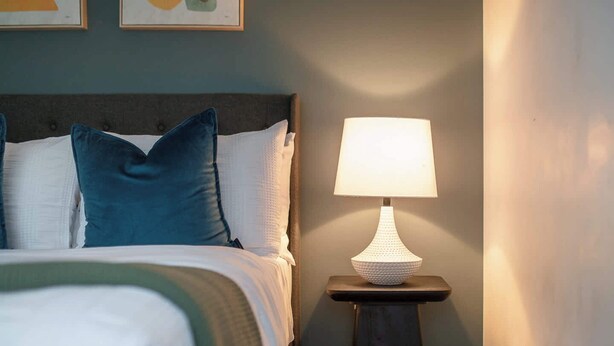 Ein ordentlich bezogenes Bett mit blau-weißen Kissen, eine weiße Lampe auf einem kleinen Tisch daneben und eine Wand mit einem gerahmten Bild.