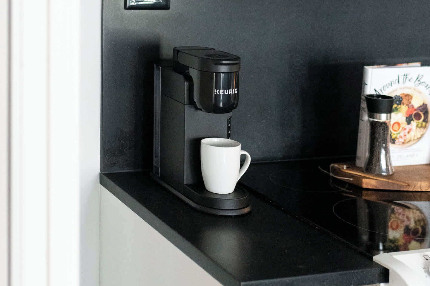 Eine Kaffeemaschine von Keurig steht auf einer schwarzen Arbeitsplatte zusammen mit einer weißen Tasse und einem Buch mit dem Titel 'Around the Bay'.