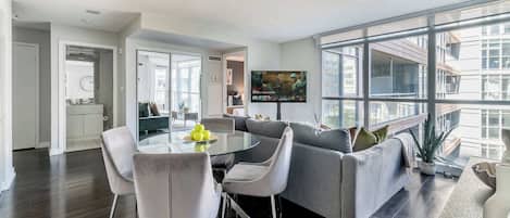 Una sala de estar moderna con una mesa de comedor de vidrio, sofás grises y un amplio ventanal con vistas a la ciudad.