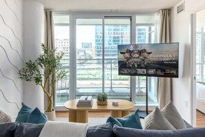 A modern living room with a large window, a flat-screen TV, a sofa, and a coffee table. - Expansive 3BR Downtown Condo with Patio (Toronto)