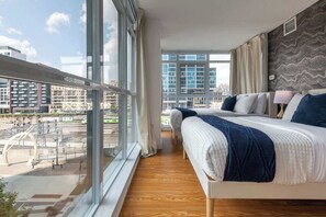 A modern hotel room with a large bed, a view of the cityscape, and a window overlooking a train station. - Expansive 3BR Downtown Condo with Patio | Parking Available (Toronto)