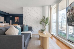 A modern living room with a grey sofa, a small round wooden table with a plant, and a large window with a city view. - Expansive 3BR Downtown Condo with Patio | Parking Available (Toronto)