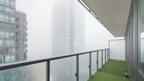 En balkon med gelænder af glas, udsigt over en by med høje bygninger, kunstgræs og et hvidt markise.