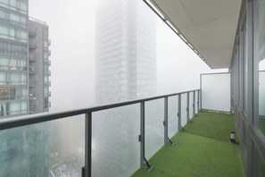Ein Balkon mit Glasgeländer und Blick auf eine Skyline mit hohen Gebäuden, einem grünen Kunstrasenteppich und einem weißen Sonnendach.