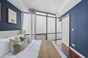 Ein Schlafzimmer mit einem großen Bett, einem Nachttisch, einer Lampe, einem Fenster mit Vorhängen und Blick auf die Stadt.