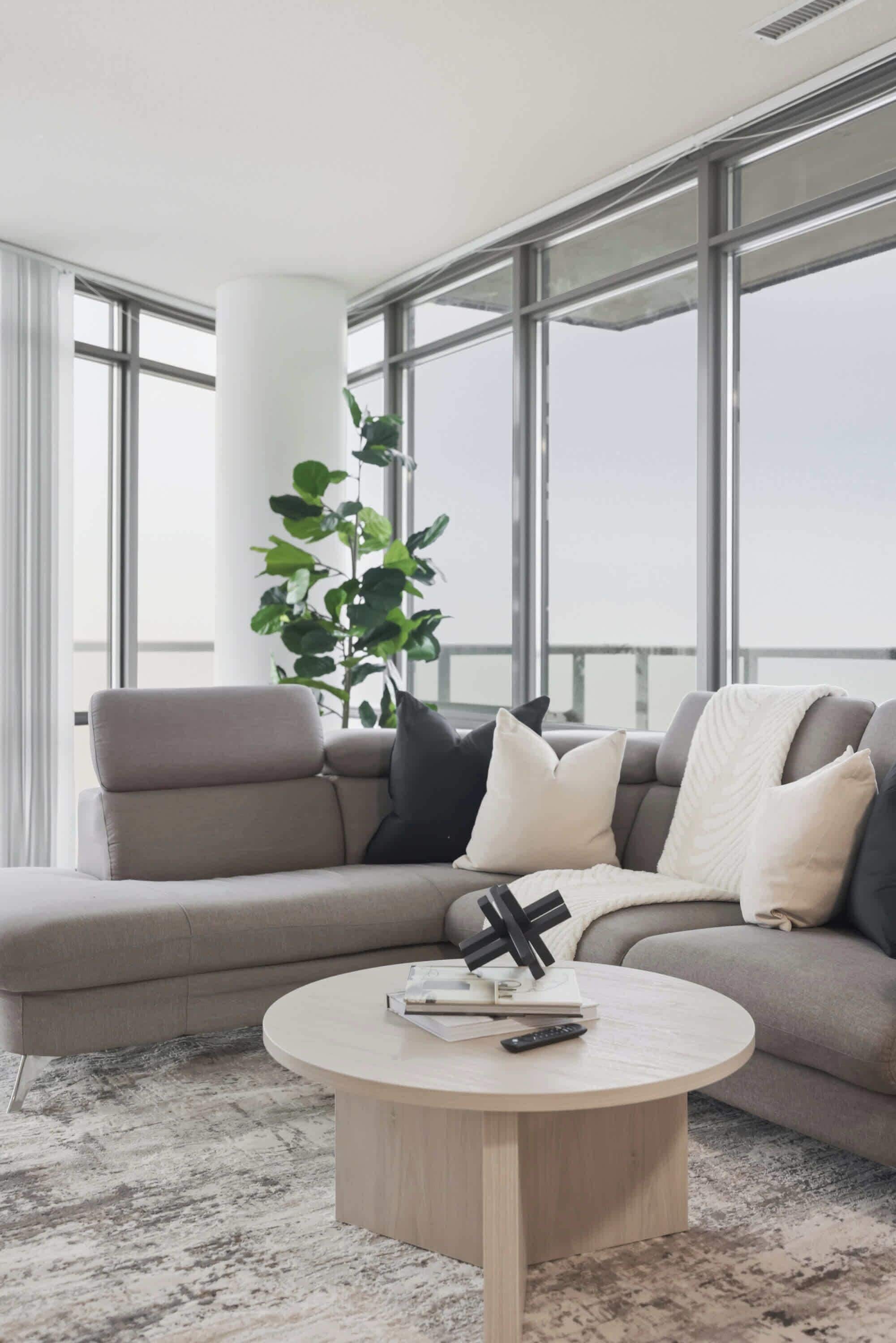Een moderne woonkamer met een grijze bank, een ronde salontafel en een groot raam met uitzicht op de lucht.