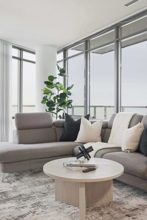 Ein modernes Wohnzimmer mit einem grauen Sofa, einem runden Couchtisch und einem großen Fenster mit Blick auf den Himmel.