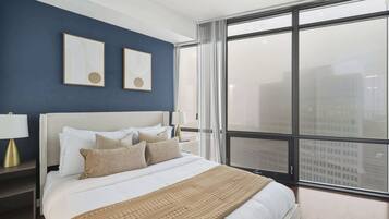 A modern bedroom with a large bed, two framed artworks on the wall, and a view of the city through floor-to-ceiling windows.