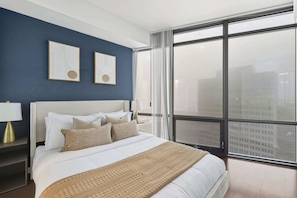 A modern bedroom with a large bed, two framed artworks on the wall, and a view of the city through floor-to-ceiling windows.