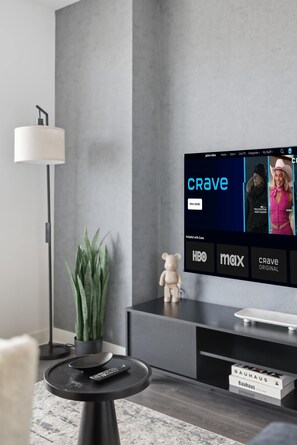 A modern living room with a flat-screen TV displaying a streaming service interface, a lamp, a plant, a remote control, and a Bauhaus-branded box. - Chic 2BR Retreat | Parking Available (Toronto)
