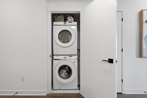 Une petite buanderie équipée de deux machines à laver superposées, d’une porte avec une poignée et d’une prise électrique murale.