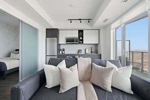 A modern living room with a grey sofa, white cushions, and a view of the city through large windows. - Chic 2BR Retreat | Parking Available (Toronto)