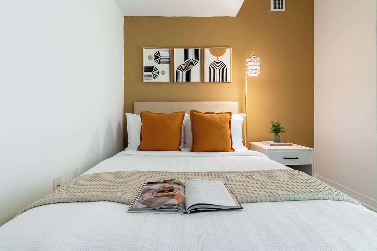 A neatly made bed with a magazine, a bedside lamp, and a nightstand with a plant.