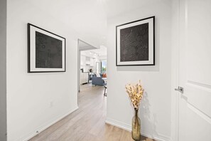 Ein Flur mit zwei gerahmten abstrakten Kunstwerken an den Wänden, einer Vase mit getrockneten Blumen auf dem Boden und einem Blick auf eine moderne Küche und ein Wohnbereich im Hintergrund.