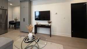 A modern living room with a flat-screen TV, a dining area with a table and chairs, and a kitchen with a refrigerator and cabinets. - Parkdale 2-Bedroom Apartment Free Parking (Toronto)