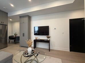 A modern living room with a flat-screen TV, a dining area with a table and chairs, and a kitchen with a refrigerator and cabinets.