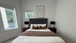 A bedroom with a large bed, two bedside tables, a headboard, and a window with blinds. - Parkdale 2-Bedroom Apartment Free Parking (Toronto)