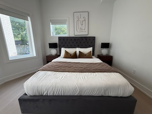 A bedroom with a large bed, two bedside tables, a headboard, and a window with blinds.