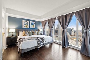 A modern bedroom with a large bed, two bedside lamps, a city view, and artwork on the wall. - Free Parking! Huge 3BR CityPlace Condo | Parking Available (Toronto)