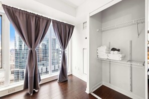 A room with a view of a cityscape through a window, a closet with white shelves, and a pair of grey curtains. - Free Parking! Huge 3BR CityPlace Condo | Parking Available (Toronto)
