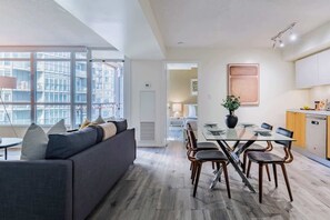 A modern living room with a sofa, dining table, and chairs, a kitchen area with cabinets and a dishwasher, and large windows offering a view of the city. - Luxury Waterfront Escape Stylish 3BR Condo | Parking Available (Toronto)