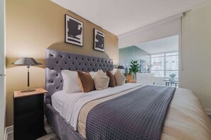 A modern bedroom with a tufted headboard, a large bed with multiple pillows, a bedside table with a lamp, and a view of the city through a large window. - Luxury Waterfront Escape Stylish 3BR Condo | Parking Available (Toronto)