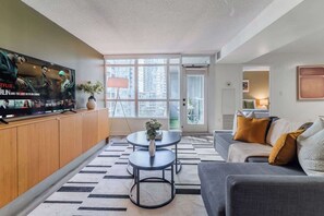 A modern living room with a flat-screen TV, a sofa, a coffee table, and a large window offering a city view. - Luxury Waterfront Escape Stylish 3BR Condo (Toronto)