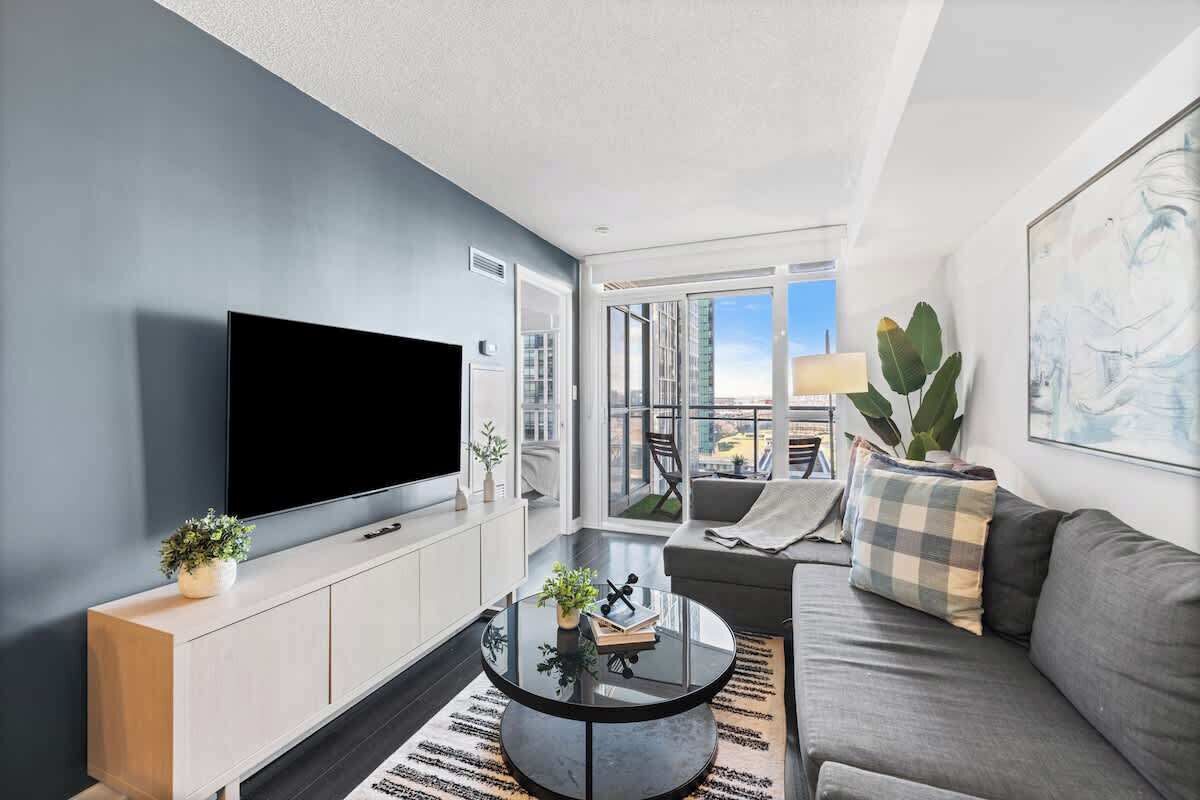 A modern living room with a flat-screen TV mounted on the wall, a round coffee table with decorative items, a grey sofa, and a large window offering a view of the city.