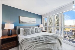 A modern bedroom with a large window, a bed with patterned bedding, a nightstand with a lamp, and a framed picture on the wall. - Downtown Central - Rogers Centre - 3 beds - Views (Toronto)