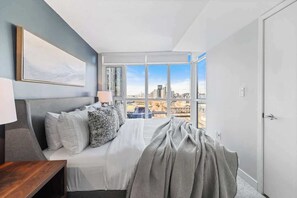 A bedroom with a bed, a nightstand, a window with a city view, and a door. - Downtown Central - Rogers Centre - 3 beds - Views (Toronto)