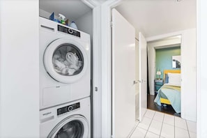 A laundry room with stacked washers and dryers, a bed in the adjacent room, and a door leading to another room. - Downtown Central - Rogers Centre - 3 beds - Views (Toronto)