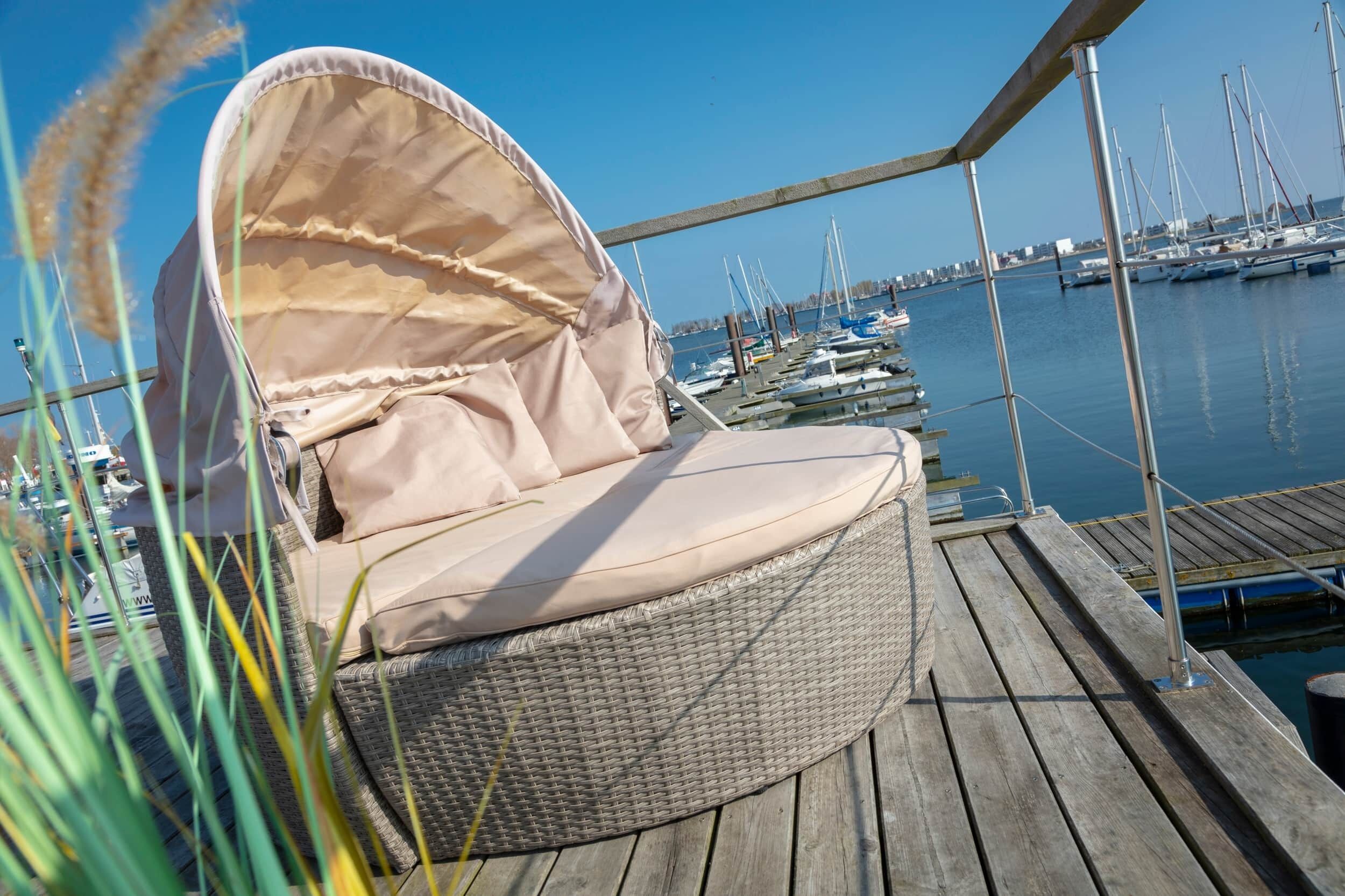 A wicker lounge chair with a canopy, positioned on a wooden deck overlooking a marina with boats.