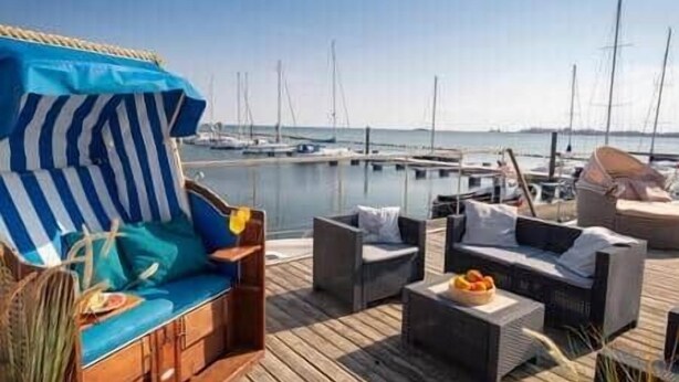 A wooden deck with outdoor furniture, including a sofa and chairs, overlooking a marina with boats. - Bosse Houseboat - Fehmarn, Burgstaaken (Fehmarn)