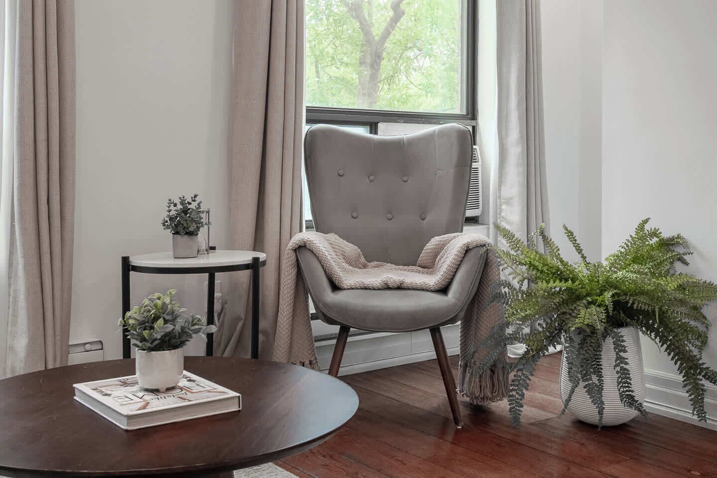 A room with a round wooden table, a grey upholstered chair, a small side table with a potted plant, and a large potted fern.