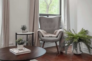 Una habitación con una mesa redonda de madera, una silla tapizada de gris, una mesita auxiliar con una planta en maceta y un helecho grande en maceta.