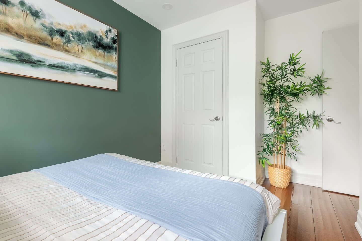 A bedroom with a bed, a door, a wall-mounted painting, and a potted plant.