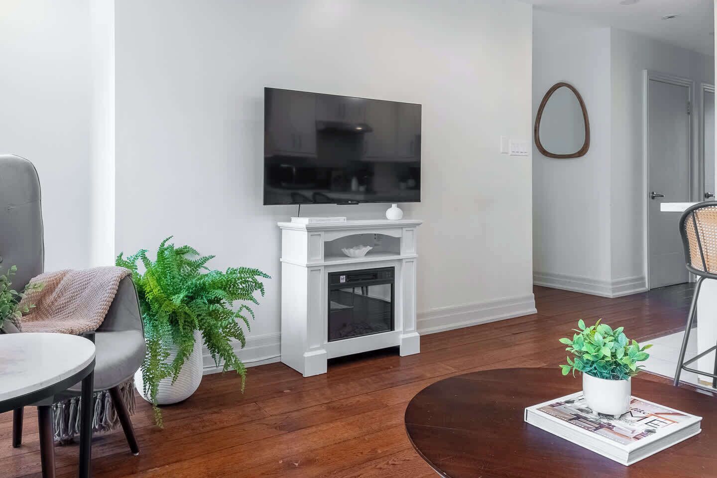 A modern living room with a flat-screen TV mounted on a white entertainment unit, a grey sofa, a round coffee table, and a potted plant.