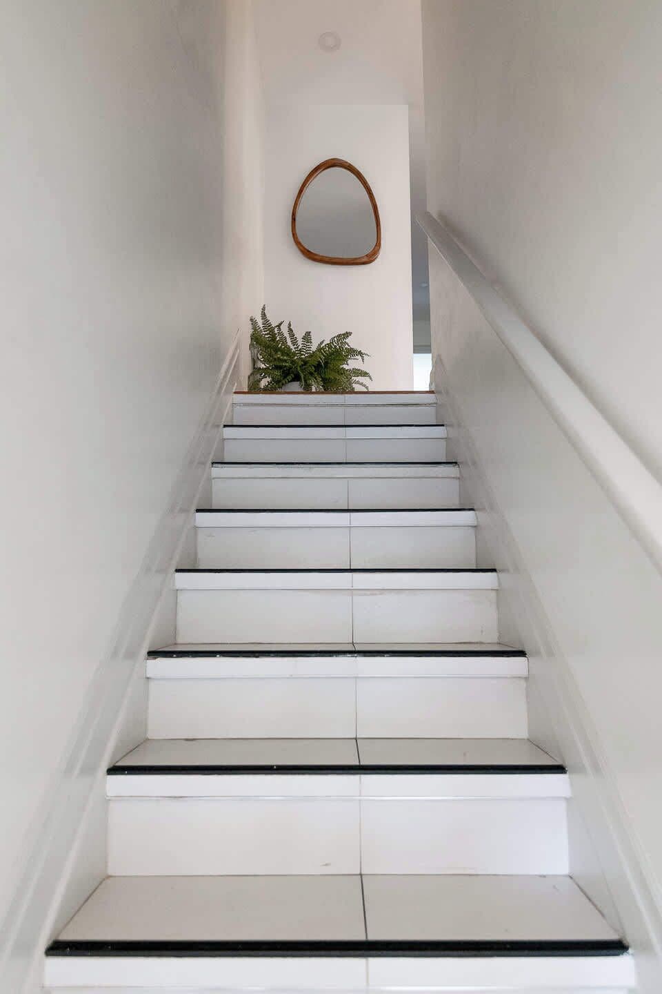 A staircase with white steps and black edges, leading to a landing with a wooden-framed mirror and a potted plant.