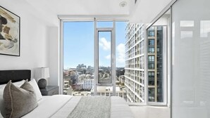 Ein modernes Schlafzimmer mit einem großen Bett, Blick auf die Stadt durch ein bodentiefes Fenster und einer sichtbaren Stadtansicht.