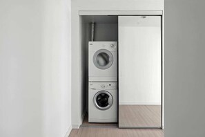 A compact laundry room with a front-loading washer and dryer, a visible hose, and a wooden floor. - Luxury 2BR Suite in Heart of Toronto Entertainment | Parking Available (Toronto)