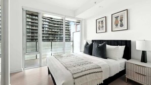 Ein Schlafzimmer mit Bett, Nachttisch, Lampe, großem Fenster und einer Glastür, die auf einen Balkon mit Blick auf Gebäude führt.