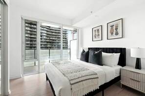 A bedroom with a bed, a nightstand, a lamp, a large window, and a sliding glass door leading to a balcony with a view of buildings. - Luxury 2BR Suite in Heart of Toronto Entertainment (Toronto)