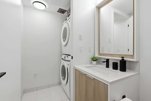 Une salle de bain moderne équipée d’un lave-linge intégré, d’un lavabo avec un miroir au-dessus et d’une petite plante en pot.