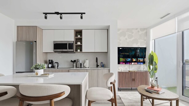 A modern kitchen with a central island, bar stools, a dining area, and a living space with a TV.