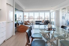 A modern living room with a glass coffee table, seating area, and large windows offering a city view. - Free Parking! Grand 3BR/2BA Suite | Sleeps 8 | Parking Available (Toronto)